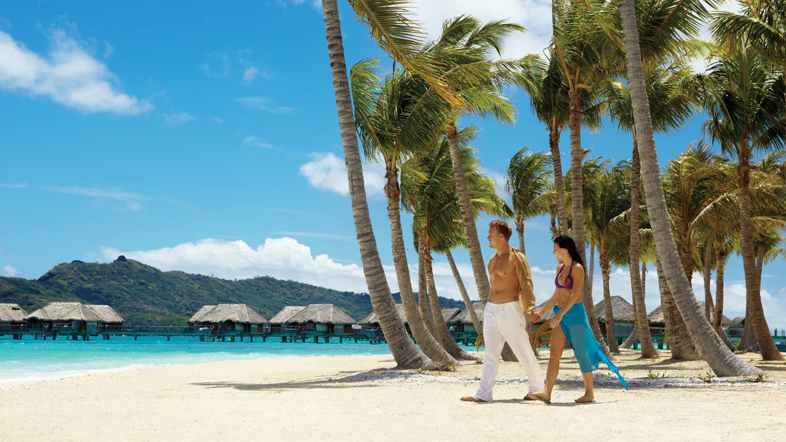 Man in white pants and woman in bikini top and blue sarong hold hands on white sand beach