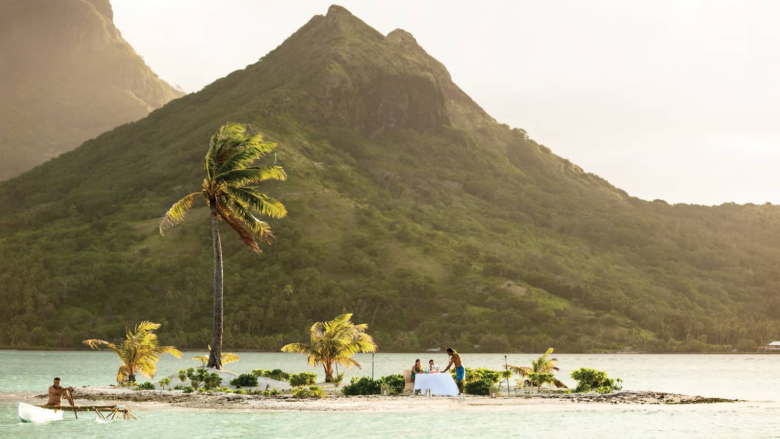 Server stands in beside couple at formal dining table on private white sand motu, lagoon and mountain behind