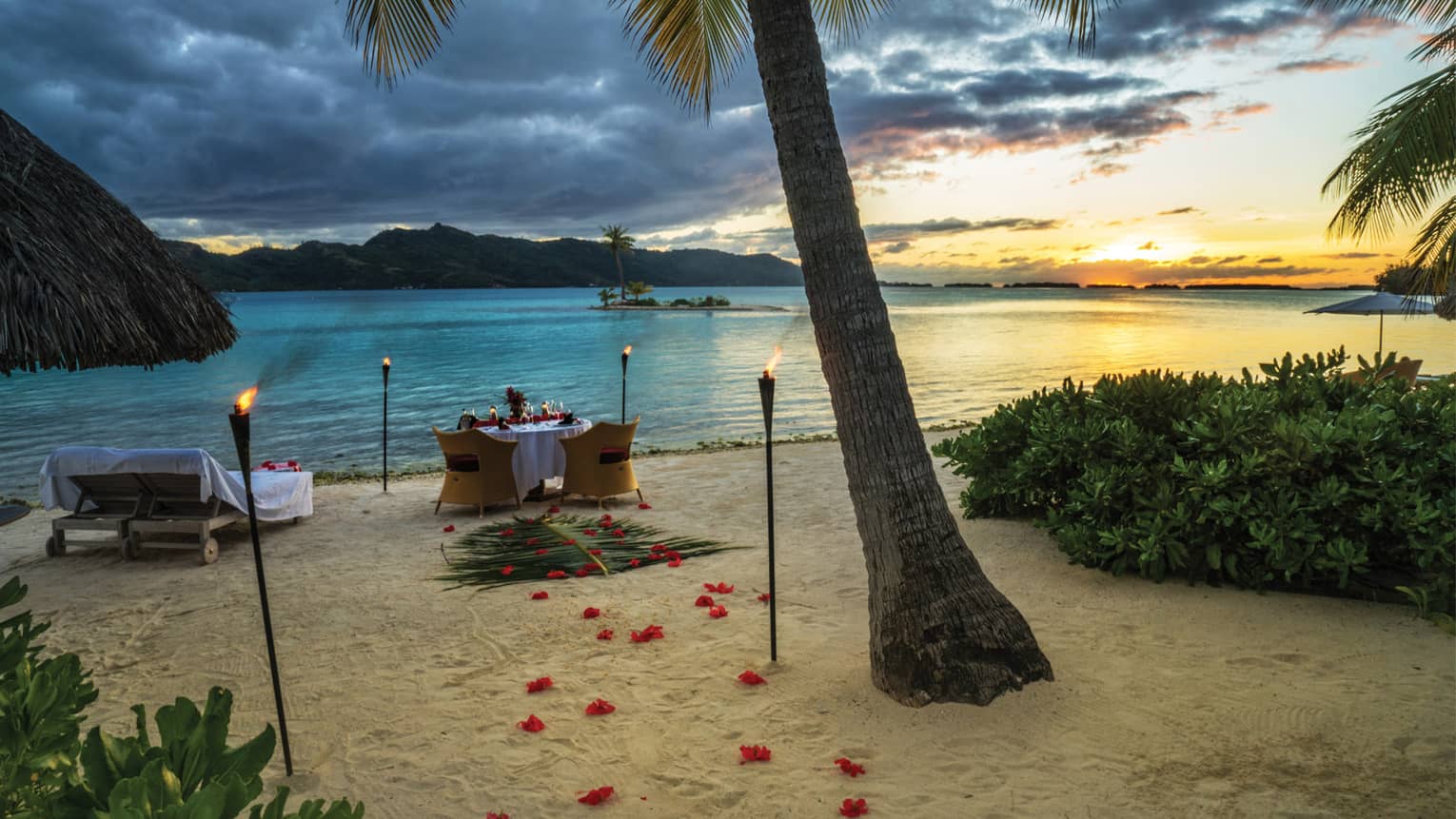 Rose petals and torches lead to a candlelit dinner table on the white sand beach by shore  at sunset