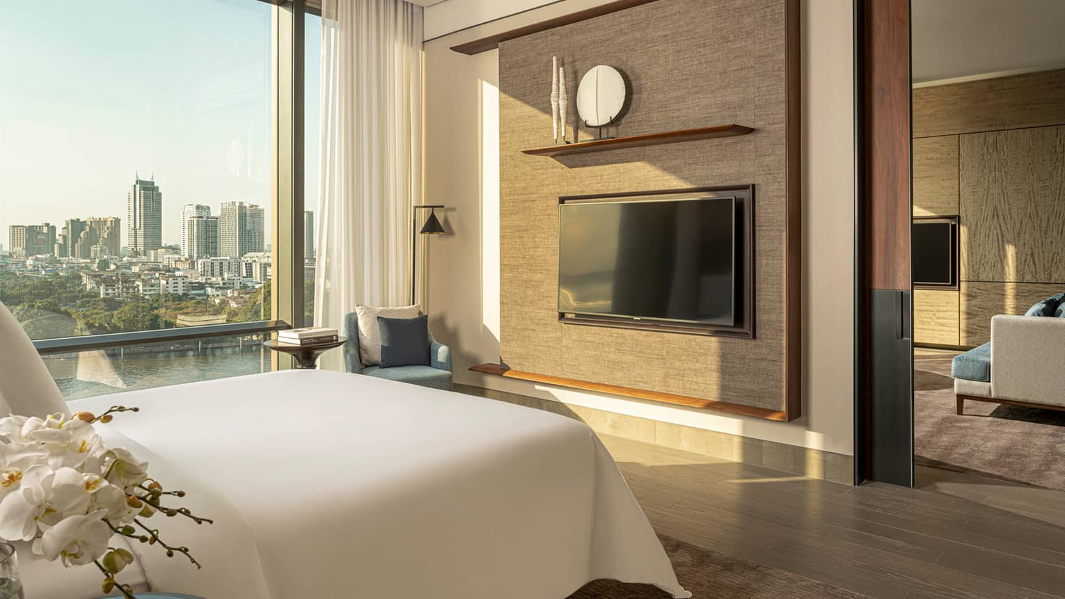 White king bed facing dark tan wall with TV, display shelf beside tall window with river and city view
