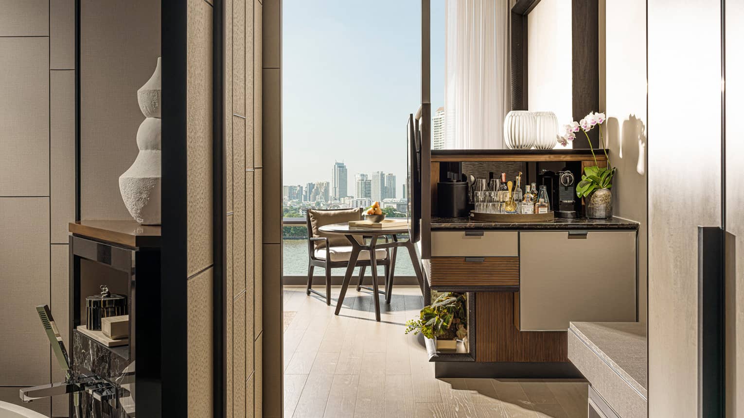 Hotel room entrance, glimpse of bathroom to left, wet bar, small dining table by tall window, city view