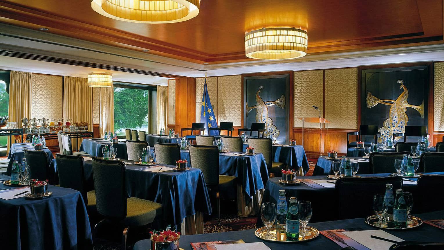 Function room with tables with blue cloths, bottles of water, facing podium 