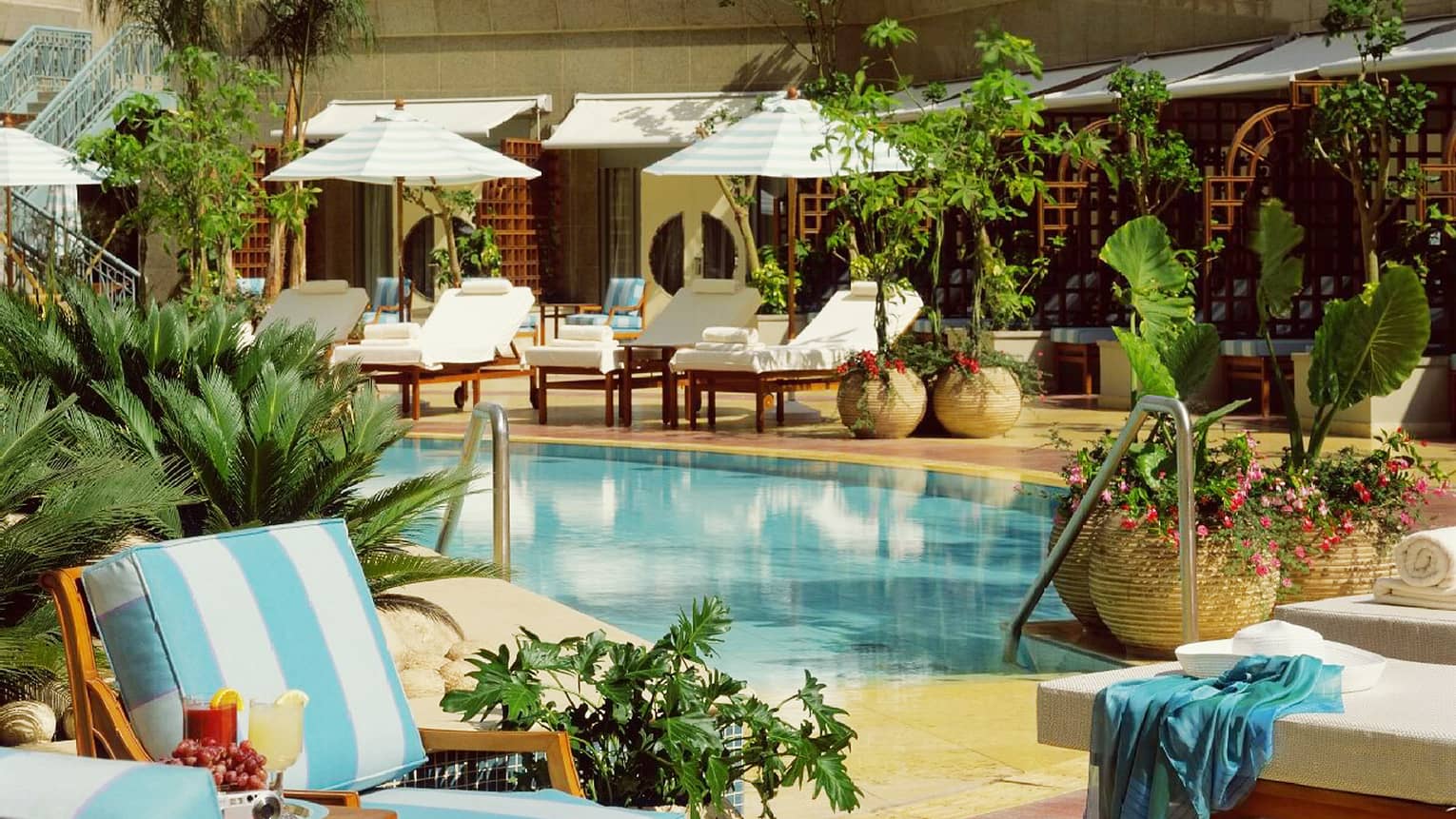 Two colourful cocktails beside blue-and-white lounge chairs, near swimming pool lounge with umbrellas, fresh flowers 