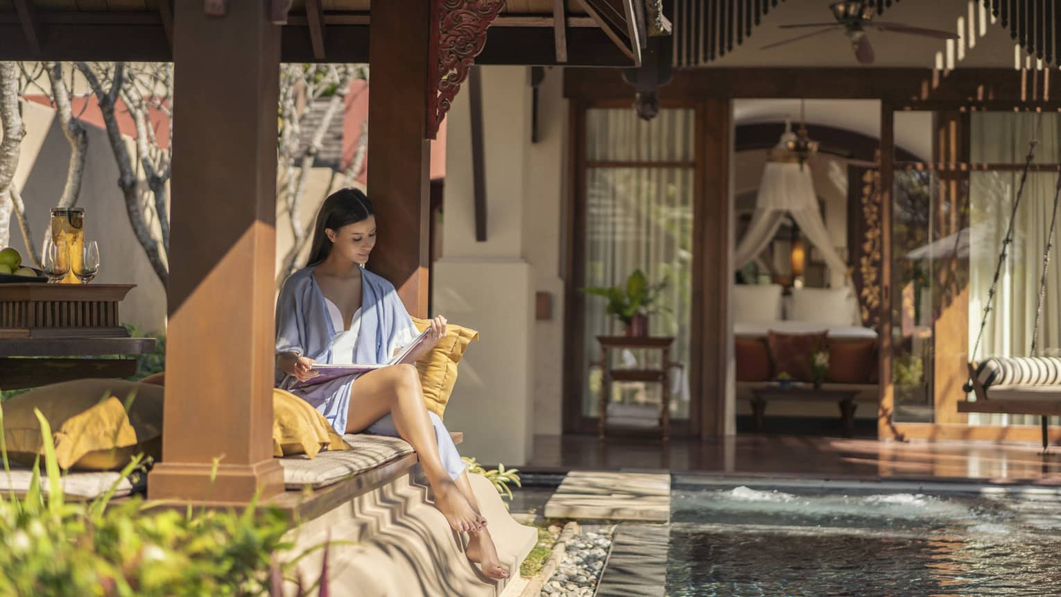 Person sitting under a shaded pavilion reading a magazine beside a private pool with a view into a bedroom featuring a canopy bed