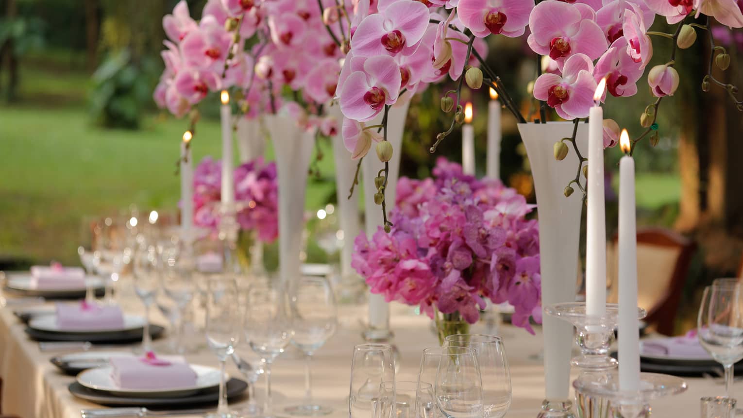 Outdoor dining table with pink flowers in vases, tall white candles 