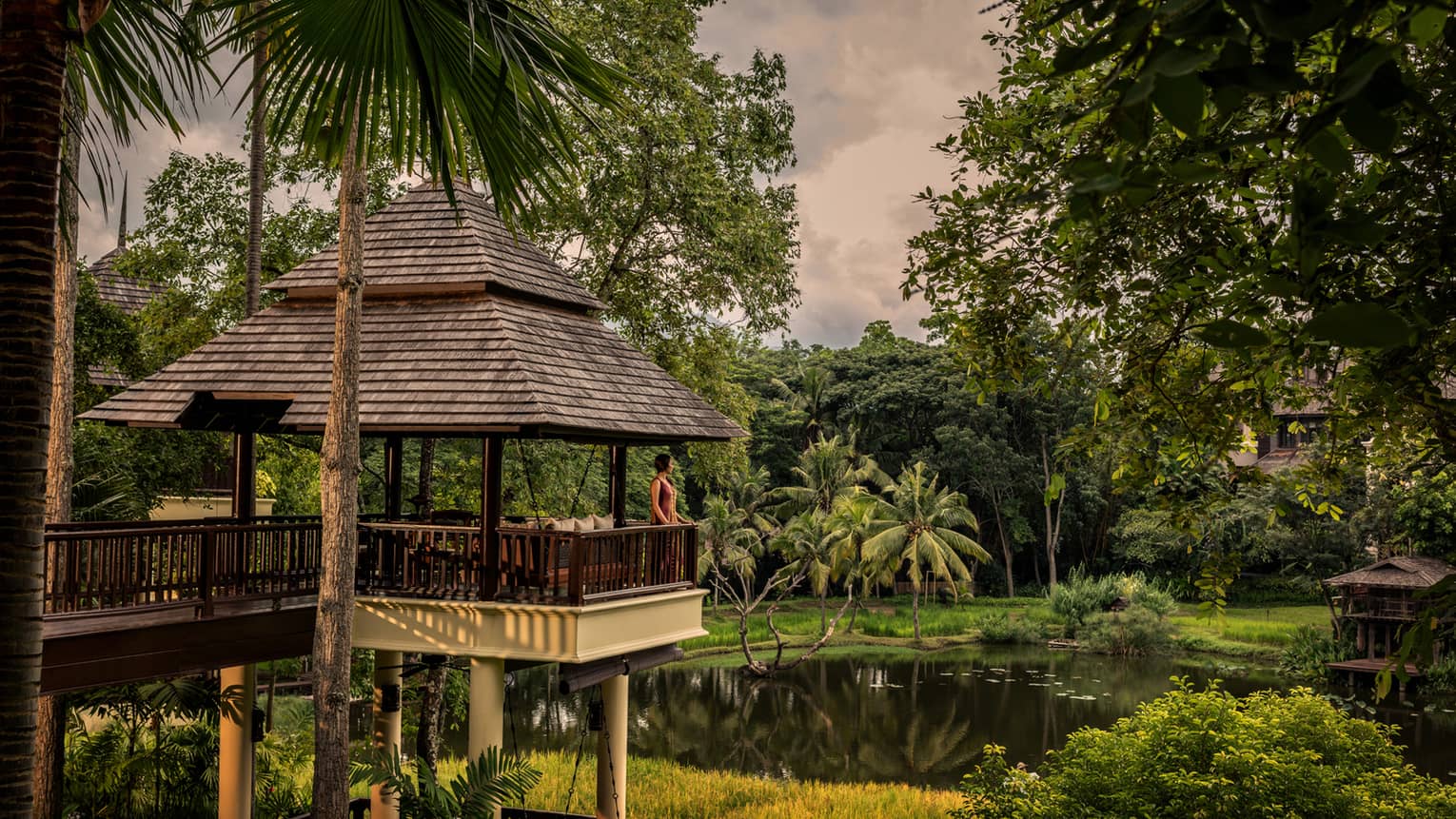 Woman stands on balcony at Upper Rice Terrace Sala overlooking tropical gardens, pond