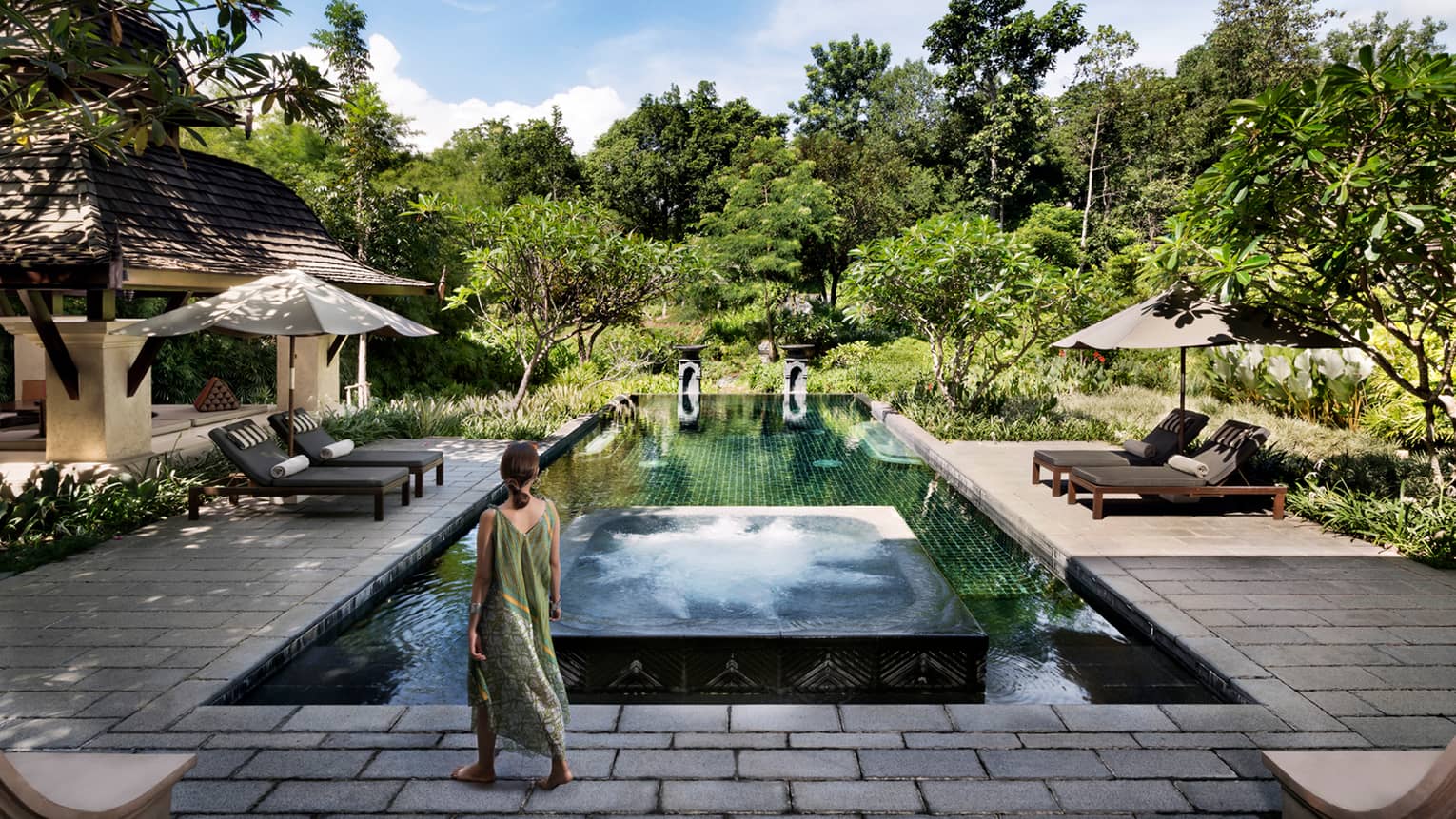 Back of woman in long beach dress walking towards residence swimming pool surrounded by tropical forest