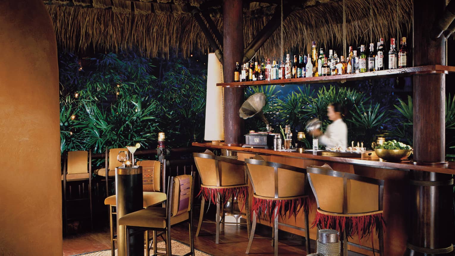 Bartender at Burma Bar with wood stools, tables under thatched-roof at night