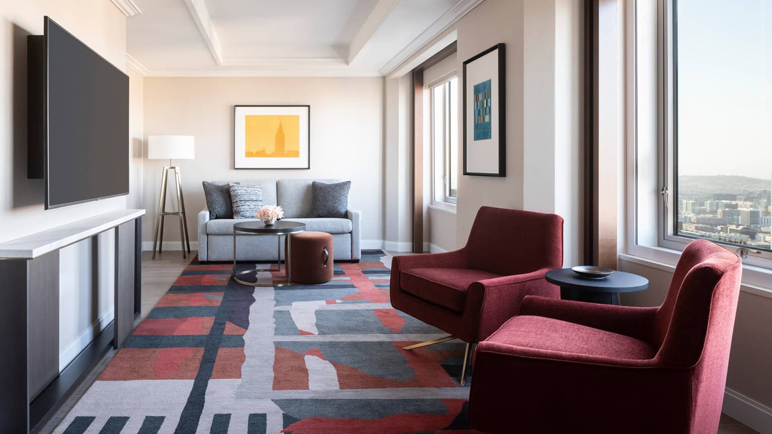 Hotel living room with two maroon arm chairs, carpet with geometric patterns, sofa, TV, windows