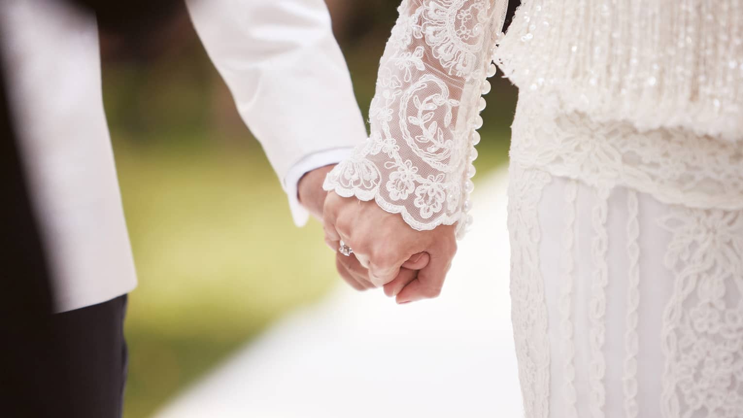 Close-up of bride with lace sleeve and groom with white suit holding hands
