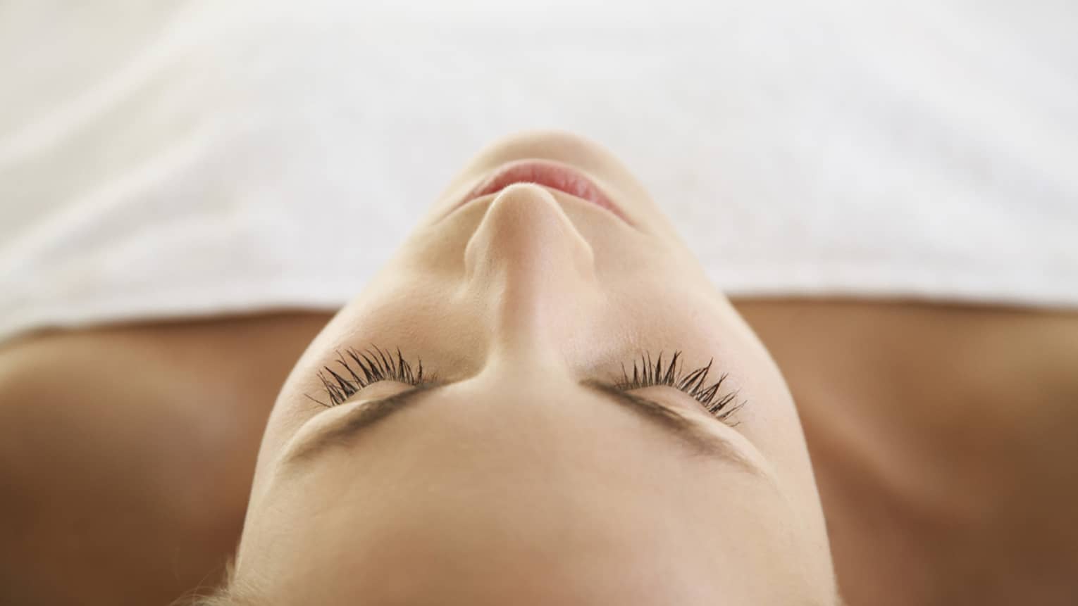 Woman lying down with eyes closed, bare shoulders, covered with white sheet