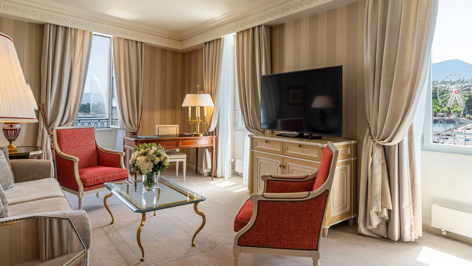 Suite living area with tall curtains, beige sofa, two red wing chairs and console with large TV