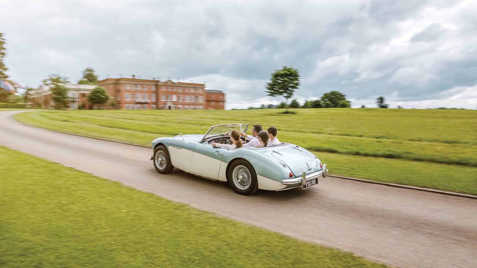 Family rides in vintage convertible car down road past English countryside to hotel manor