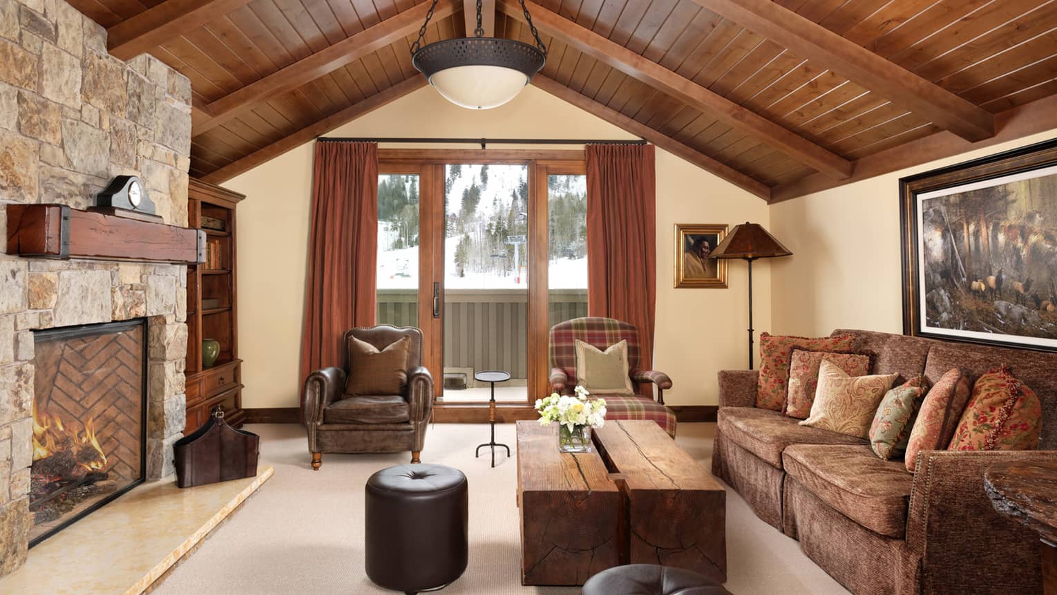 Living room with vaulted wood ceiling, stone fireplace, wood coffee table, brown sofa, leather armchair and ottomans, and plaid accent armchair