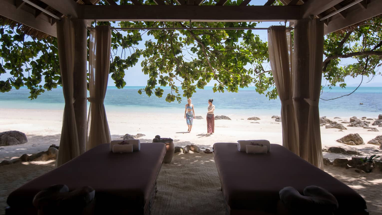 Couples massage beds under cabana in front of two women walking on white sand beach