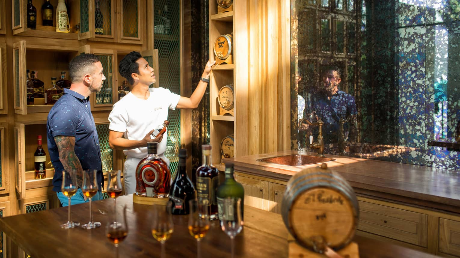 Two men examine rum barrell on shelf in wood liquor cellar, bar with bottles and tasting glasses