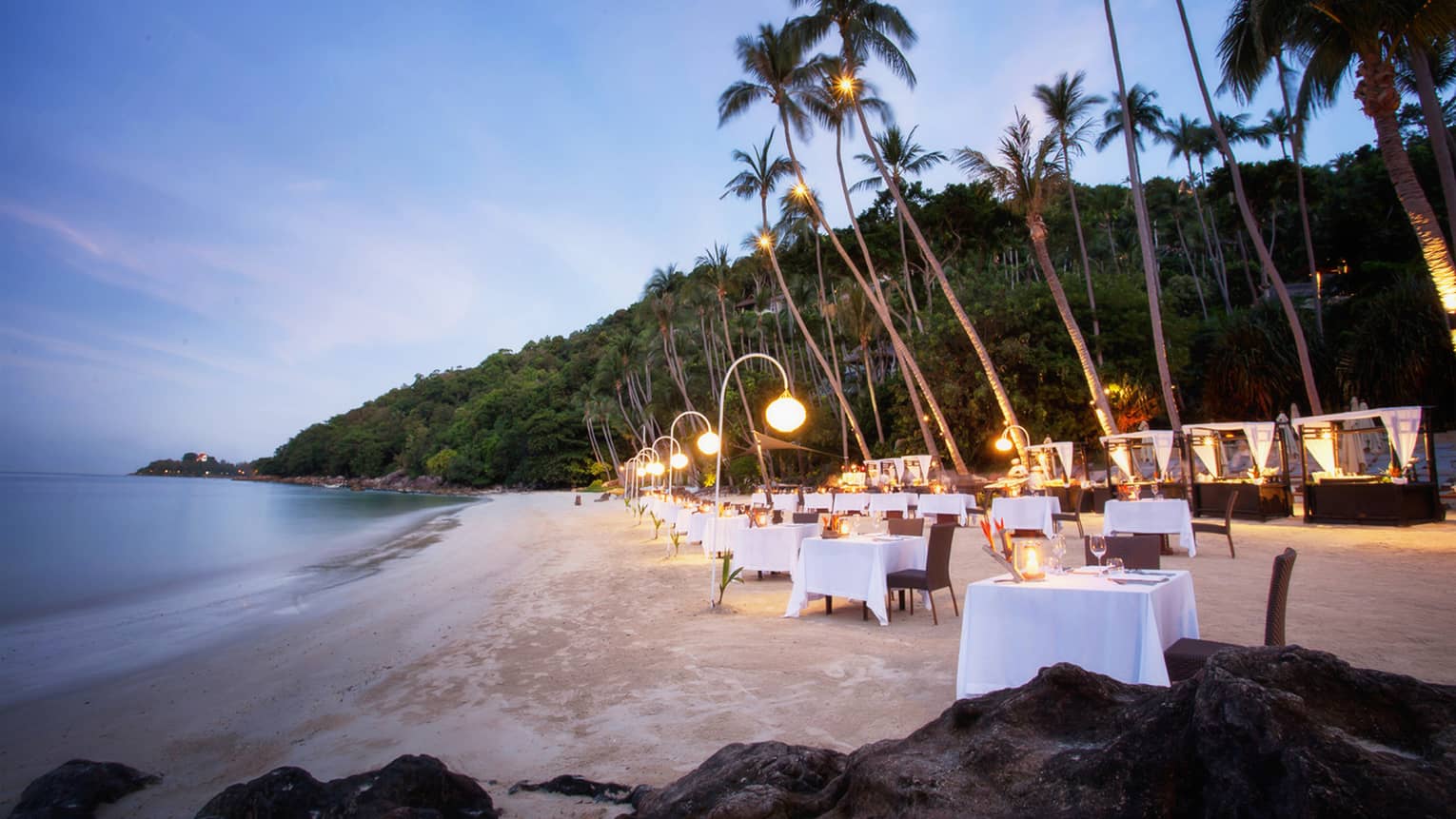 Dining tables set up along white sand beach under palm trees at dusk