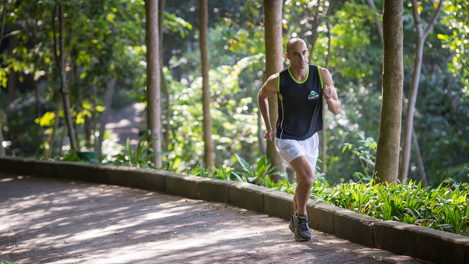 Man in workout shirt, shorts runs up pathway by trees on sunny day