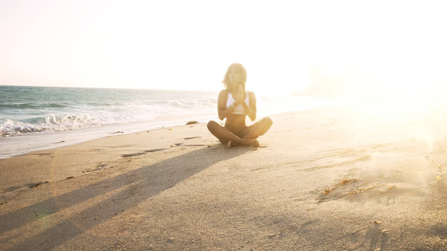 Woman in swimsuit sits cross-legged on sand beach at sunset