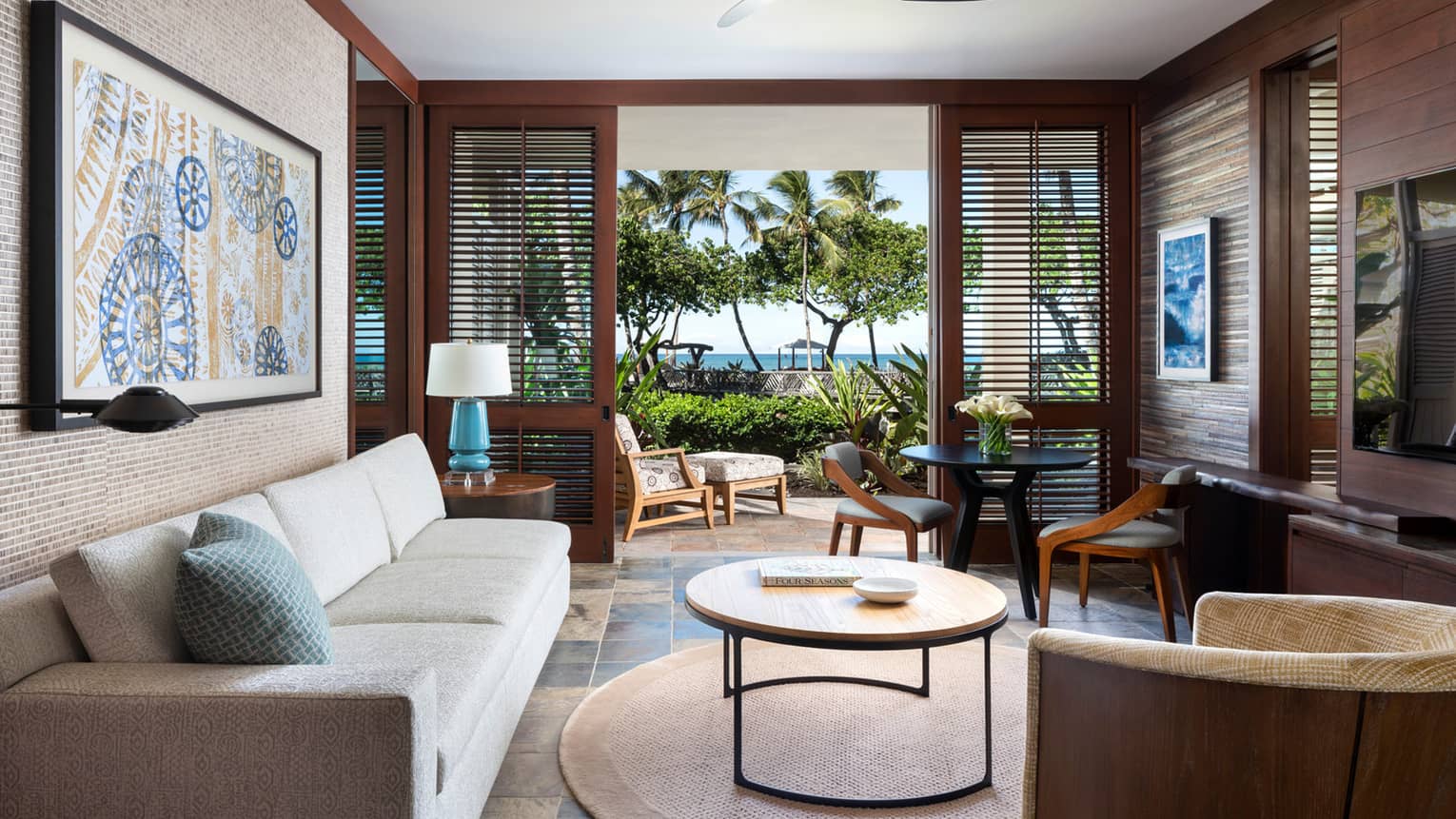 Living room with white sofa, circular coffee table, wall art, dark wooden shutter doors opening to lanai
