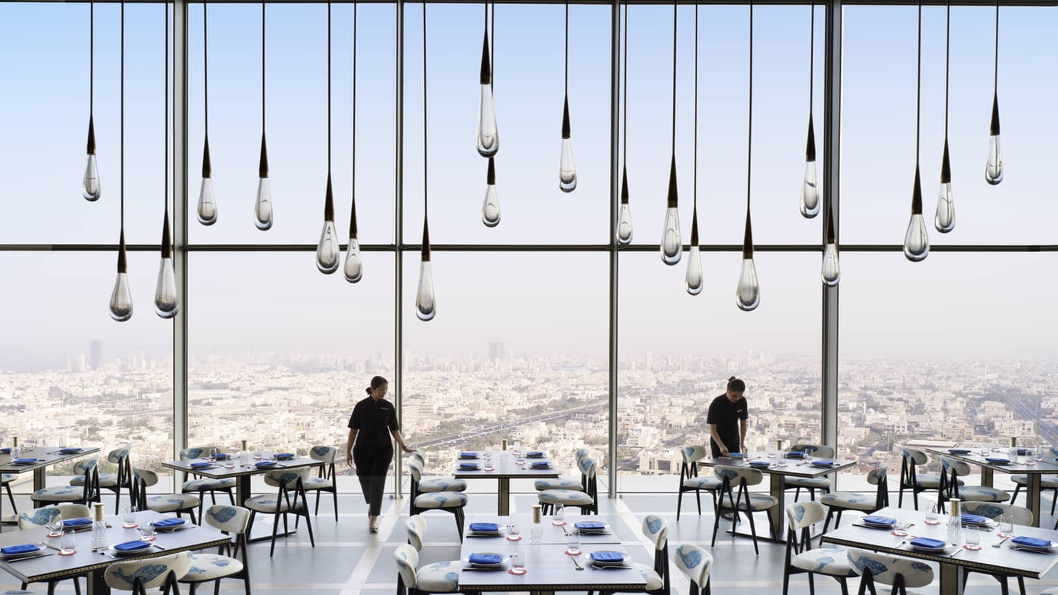 Large glass raindrop-shaped lights hang from soaring ceiling in sunny dining room, servers set tables