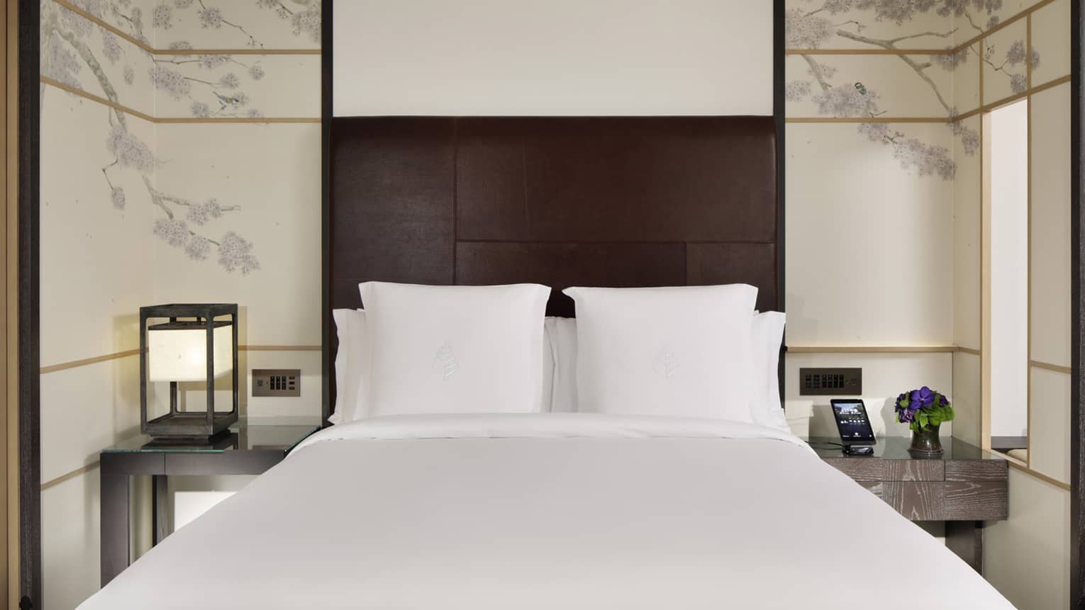 Deluxe guest room with king bed, brown headboard, decorative floral wall panels and bedside tables with a lantern lamp and flower vase
