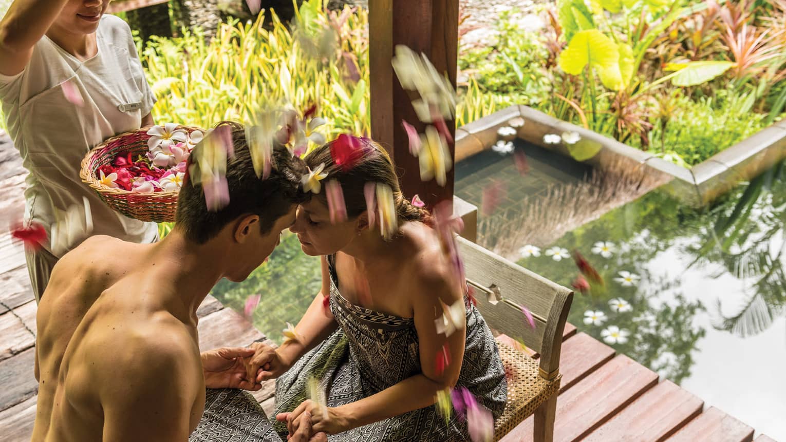 Couple on chairs, hold hands, foreheads together as woman sprinkles flower petals