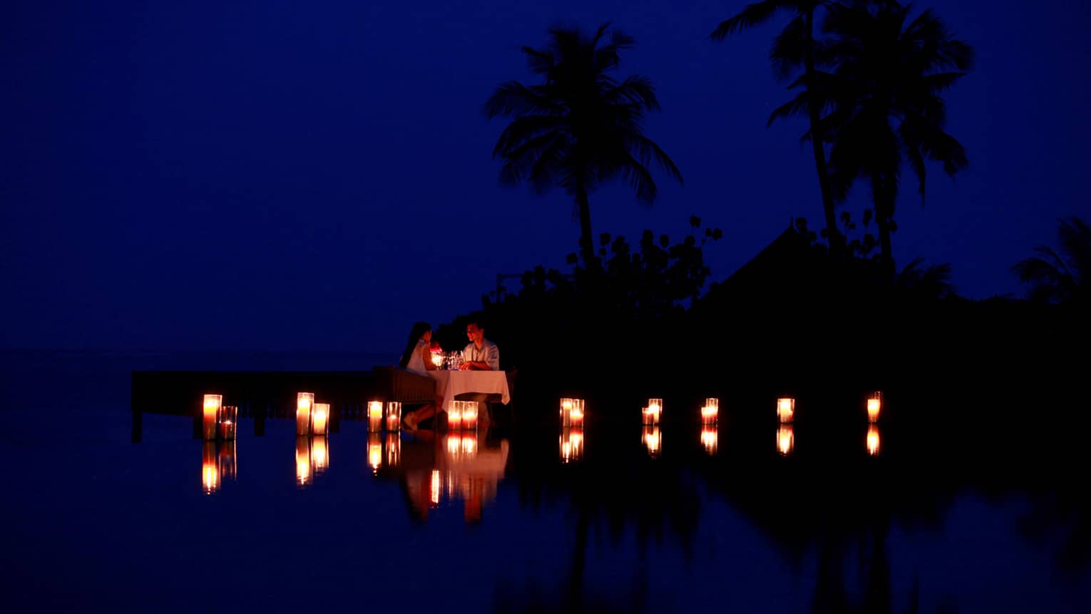 Couple dine by lagoon at small table surrounded by glowing candles at night