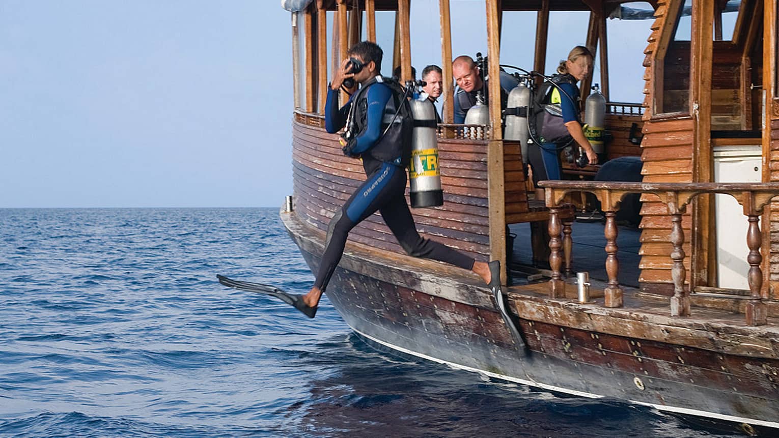 Man in SCUBA gear leaps from wooden boat into ocean and people on boat