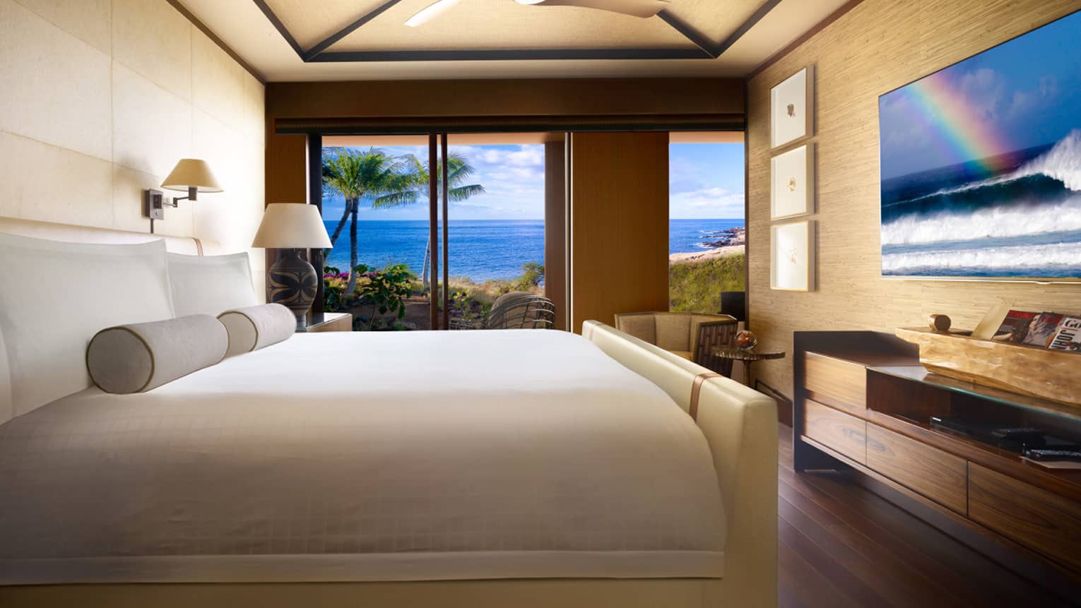 Oceanfront studio suite white bed with rolled accent pillows, modern wood dresser, flat screen TV with ocean, rainbow