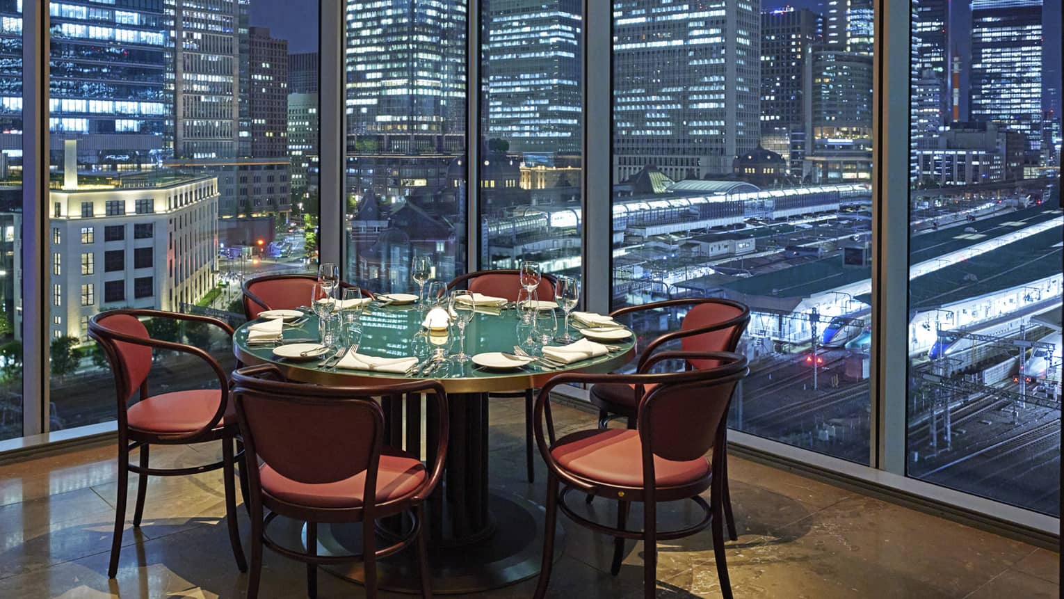 Luxury boutique hotel dining setup with a round table elegantly set for guests, offering a panoramic city view at night through tall glass windows