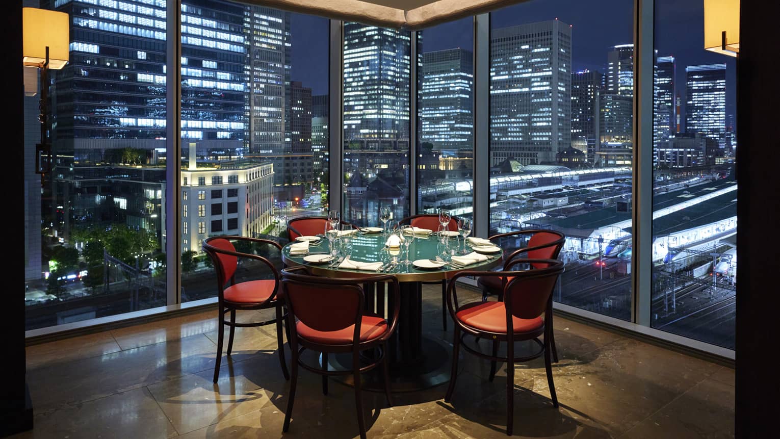 Luxury boutique hotel dining setup with a round table elegantly set for guests, offering a panoramic city view at night through tall glass windows