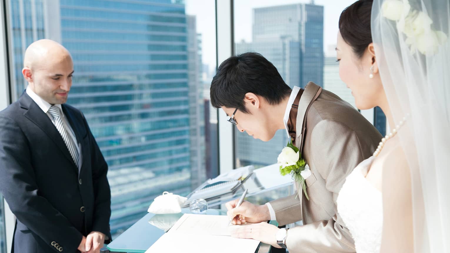 Groom signs wedding register next to bride, man wearing suit at sunny window