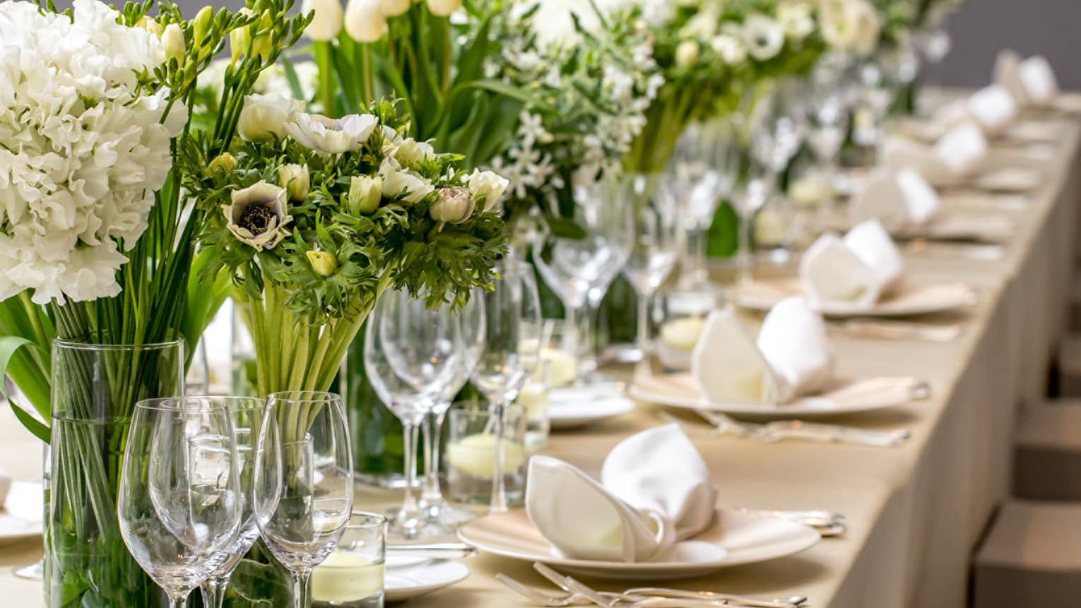 A long wedding dining table featuring white flowers in vases, and neatly organized place settings and wine glasses.