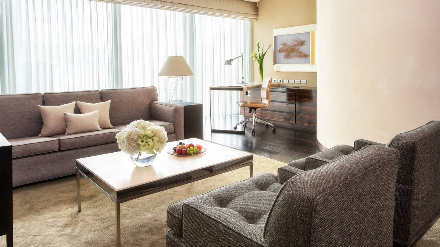Living area with grey sofas, coffee table with flowers and fruit, work desk with a chair and floor-to-ceiling windows with curtains