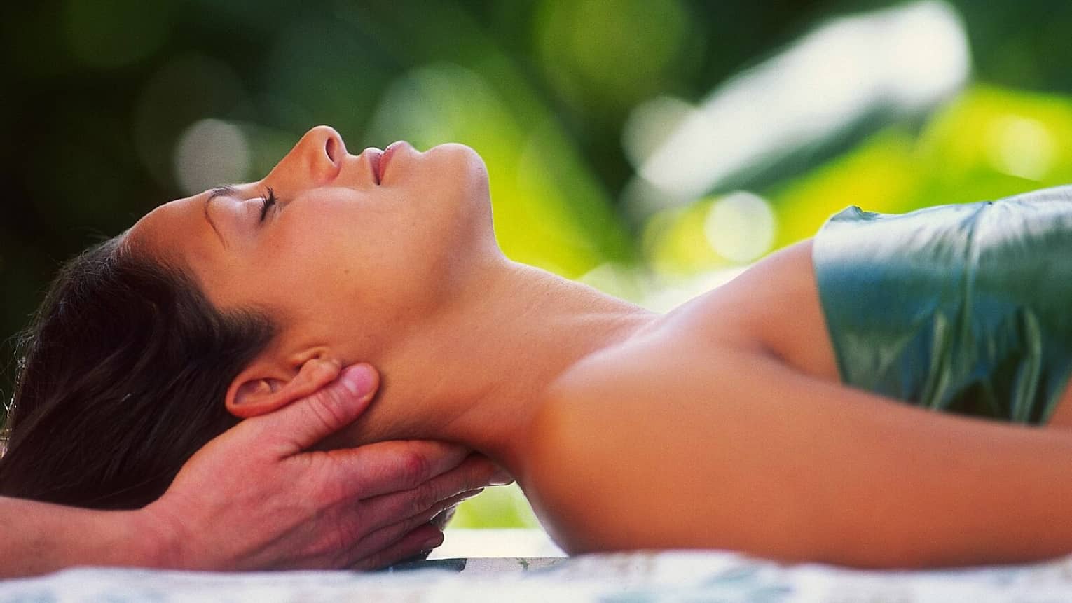 Woman with eyes closed lays on back on table with leaf over chest, hands massage her neck