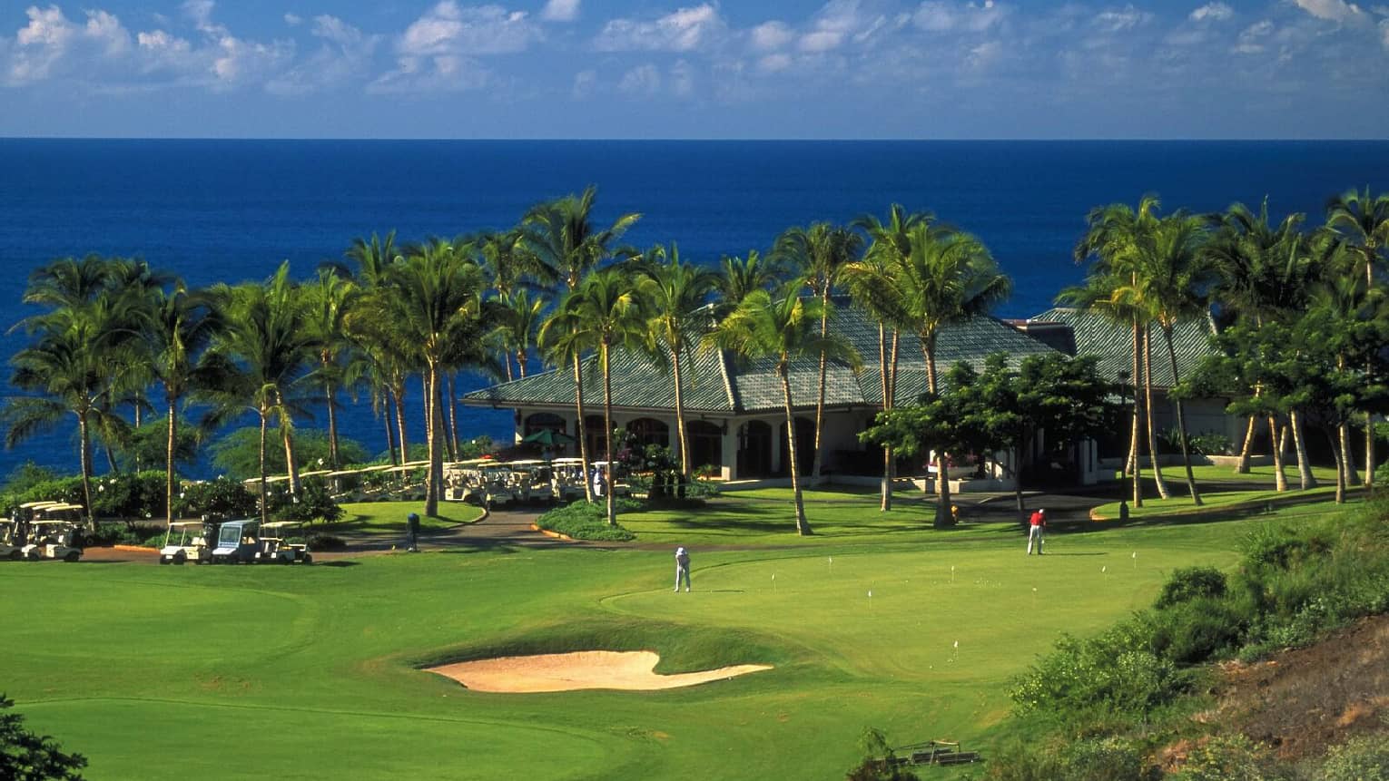 A guest plays golf on a sunny green. Palm trees are in front of a low-roofed clubhouse with the ocean in the background.