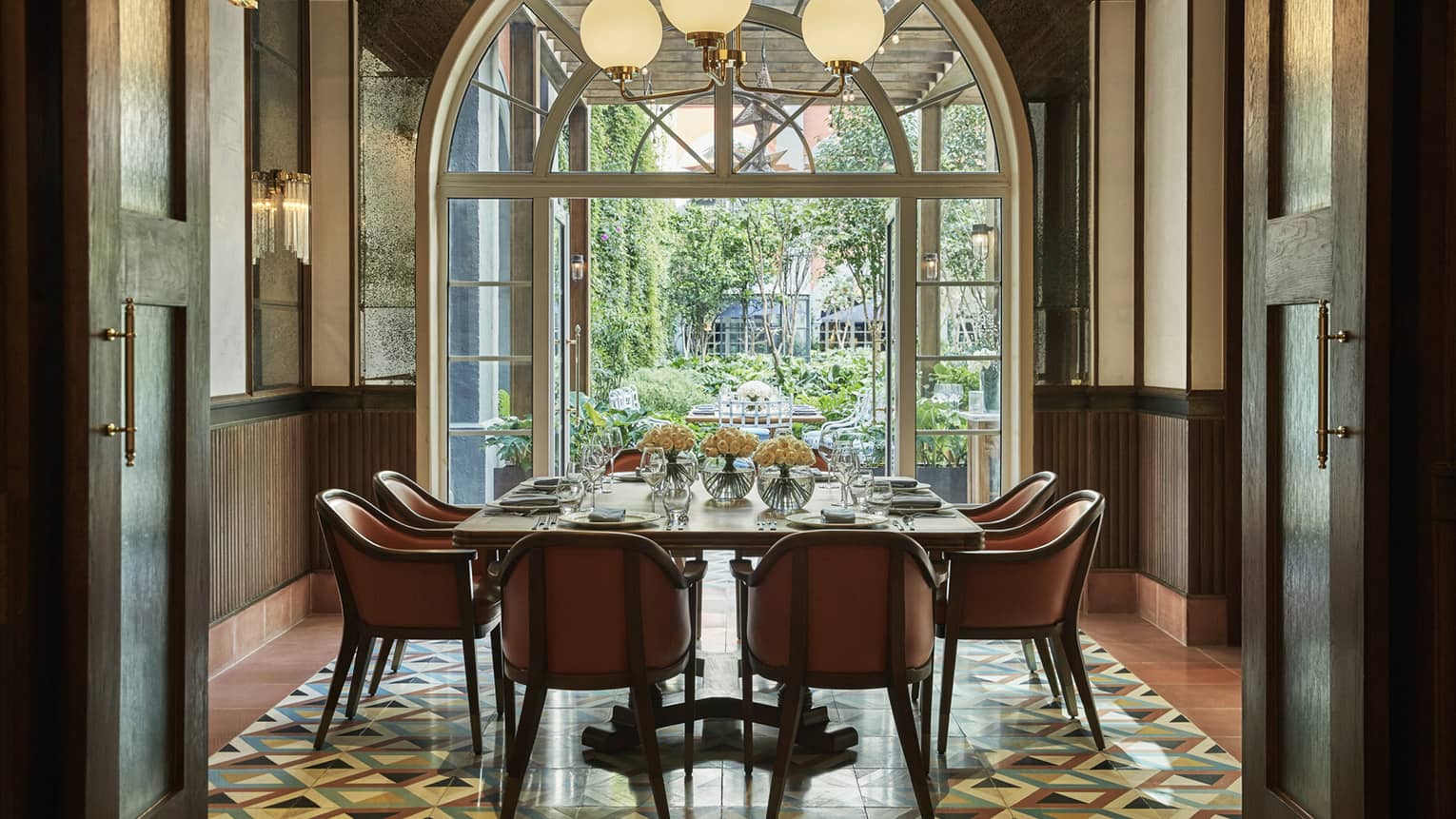 Dining table with orange chairs under globe lights by tall arched glass doors