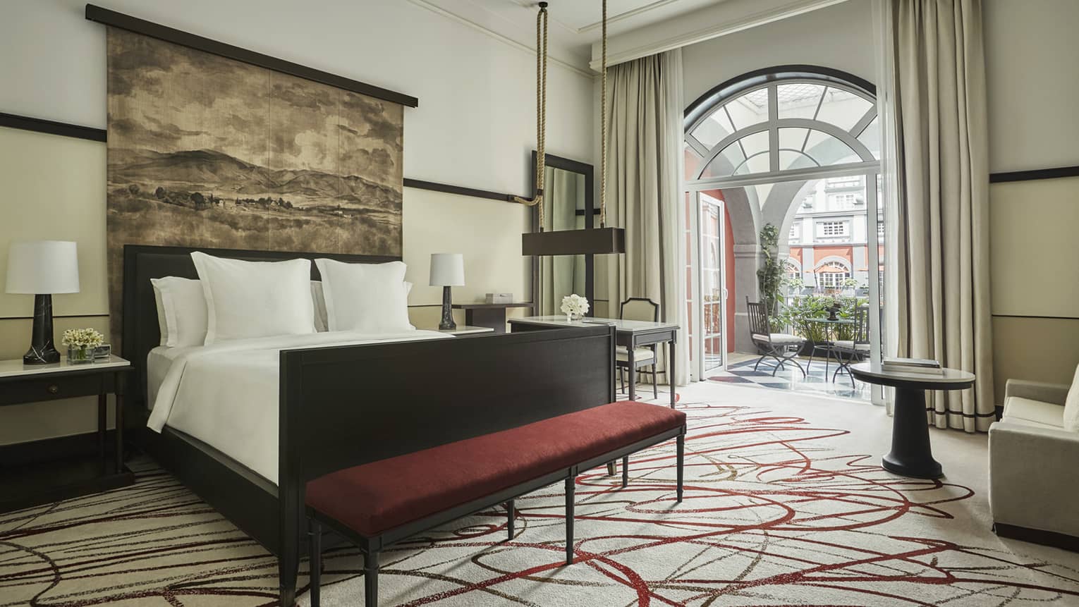 Hotel room bed under large tapestry, red velvet bench at foot, arched patio door