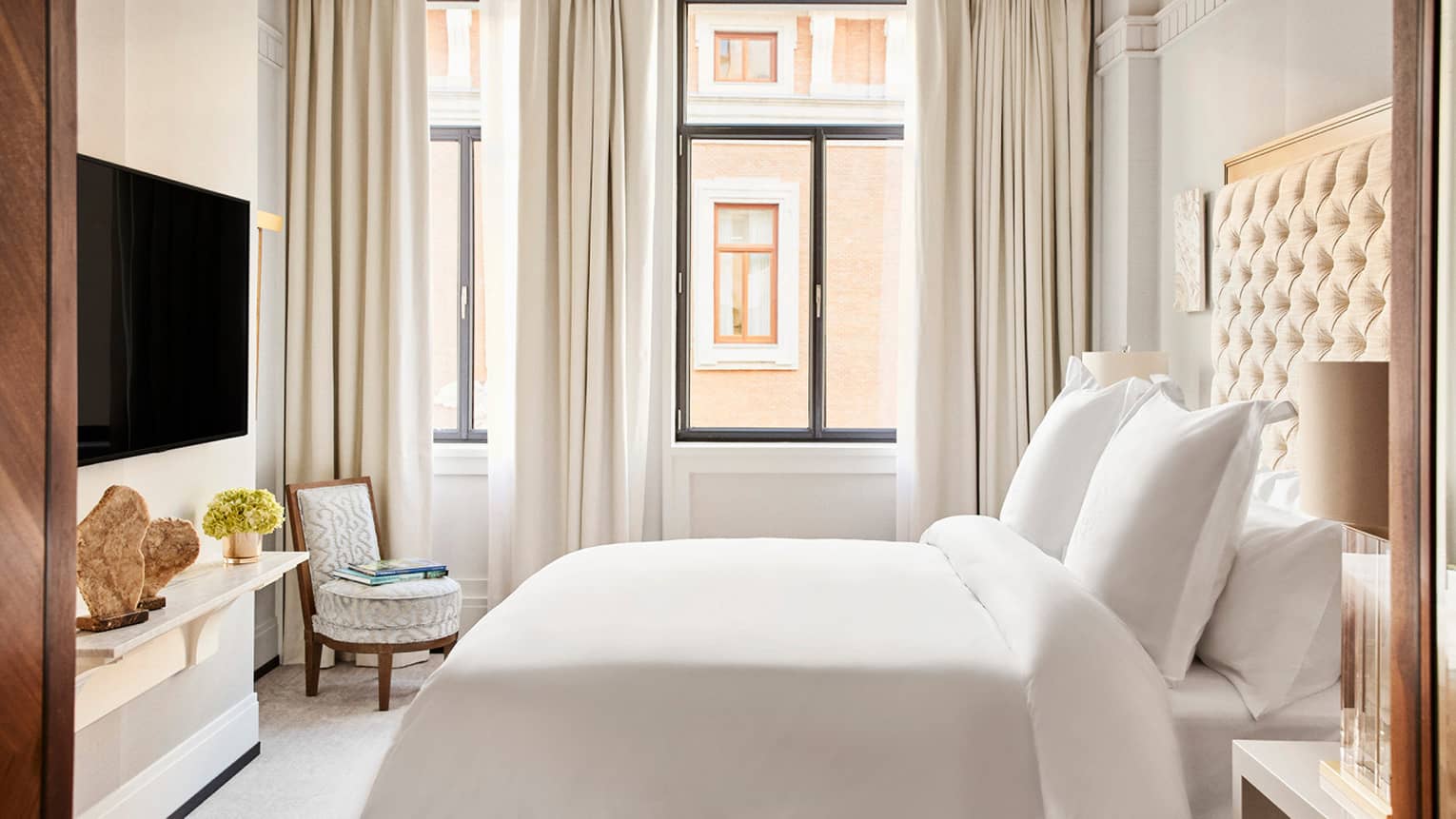 Luxury hotel suite bedroom with a king bed with tufted headboard, wall-mounted TV, small shelf with decorative stone sculptures and flowers and armchair 