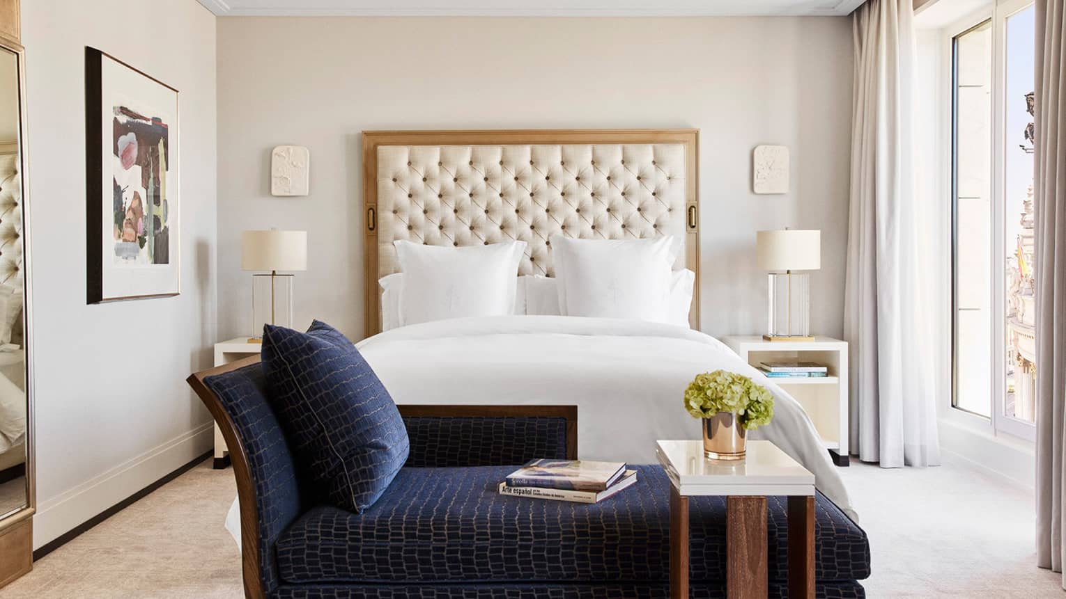 Luxury hotel bedroom with king bed, tufted headboard, blue chaise lounge with books, bedside tables with lamps, wall art and large windows 