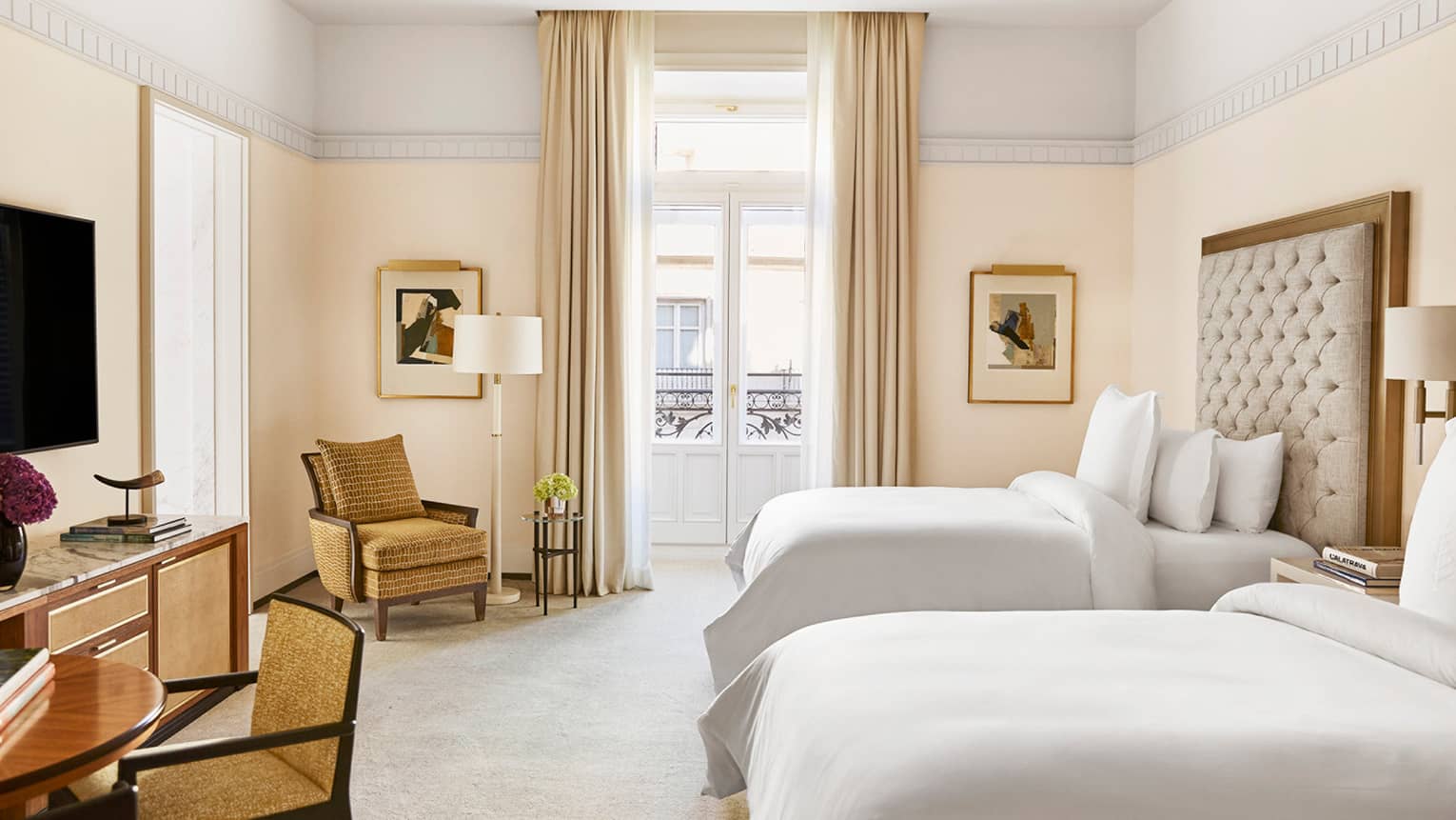 Hotel room with two beds, tufted headboard, tan armchair, round table with books, TV, dresser, wall art and French doors with curtains opening to a balcony
