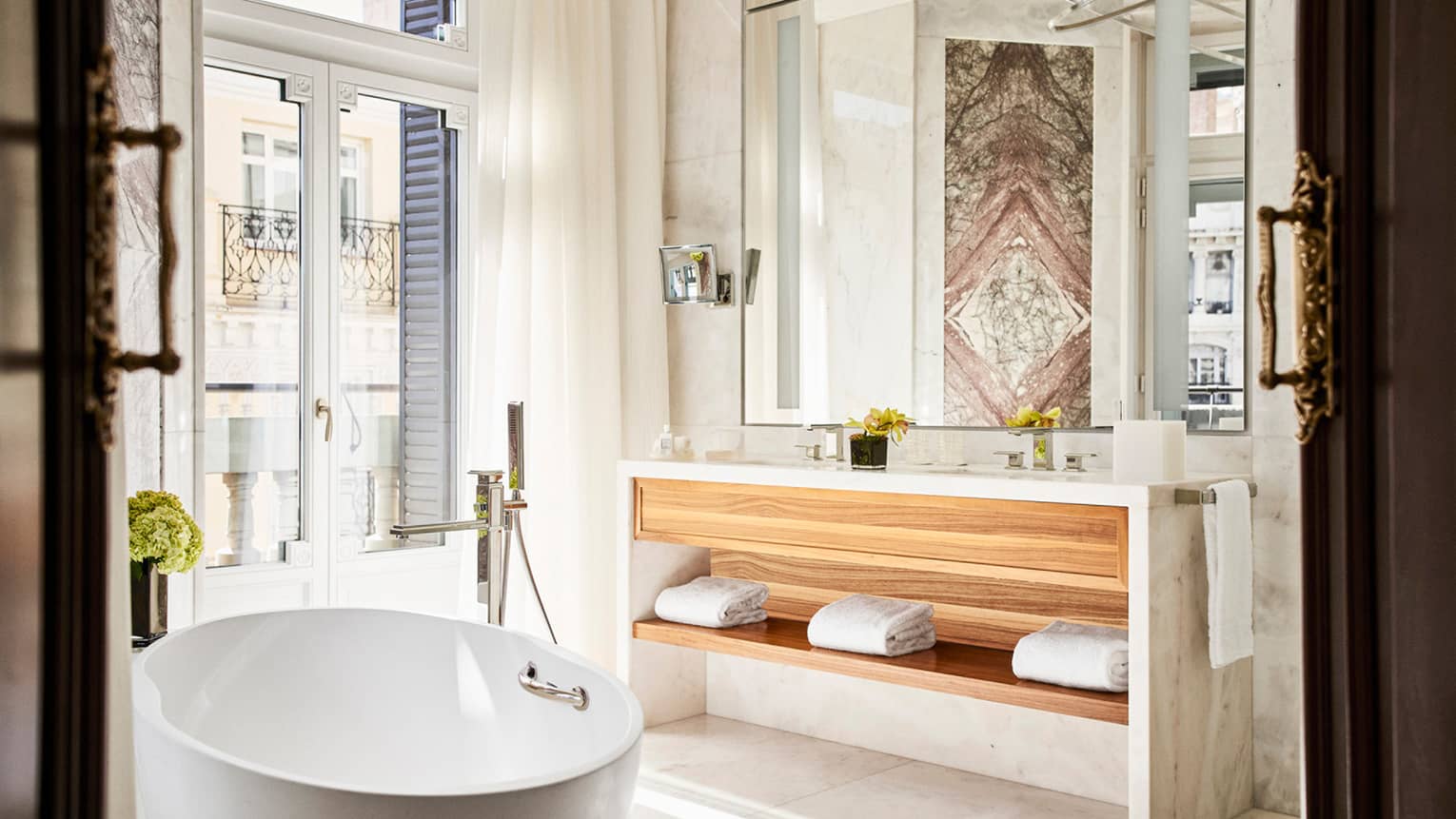 Luxury hotel bathroom with a freestanding bathtub, double vanity with wooden shelving, large mirror, marble walls and doors with balcony view 