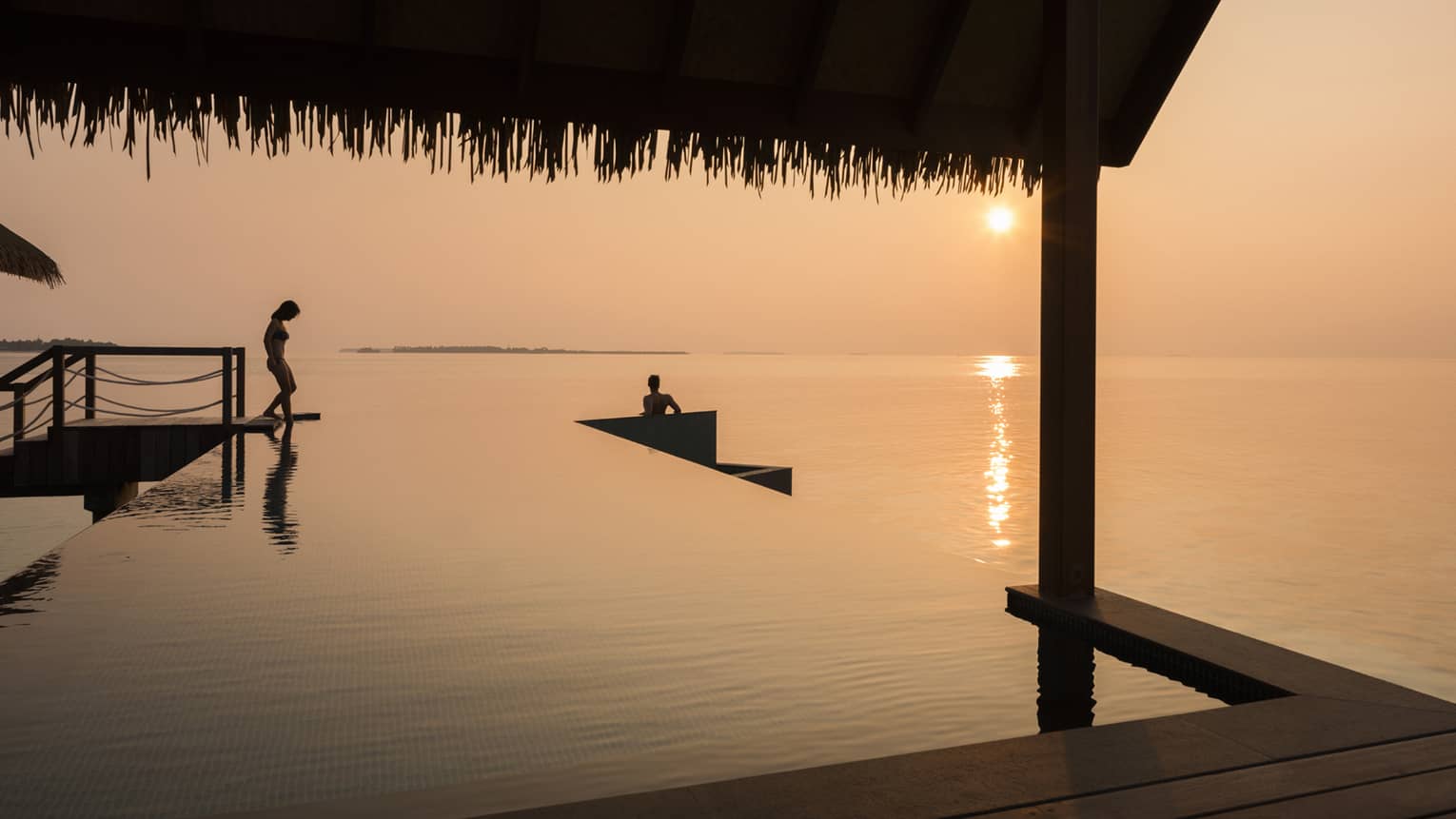 Orange sunset over ocean, silhouettes of people stepping into infinity pool under cabana