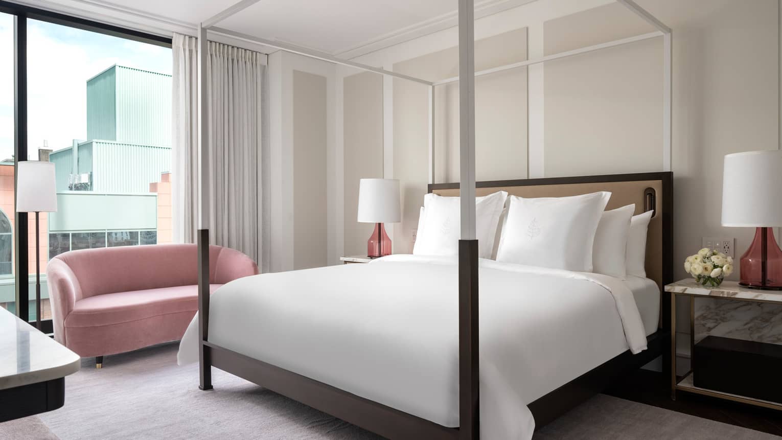 A modern hotel bedroom houses a four-poster bed, white linens and pink accent lamps. A pink loveseat sits near large windows, offering views of adjacent buildings.