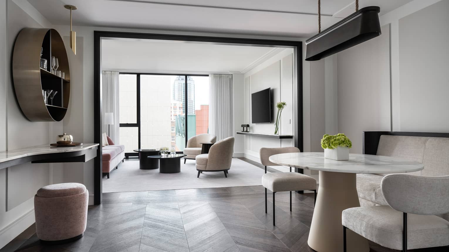 A modern living and dining space with sleek, minimalist furniture, including a round marble dining table, neutral chairs, and a lounge area with a large TV and city views