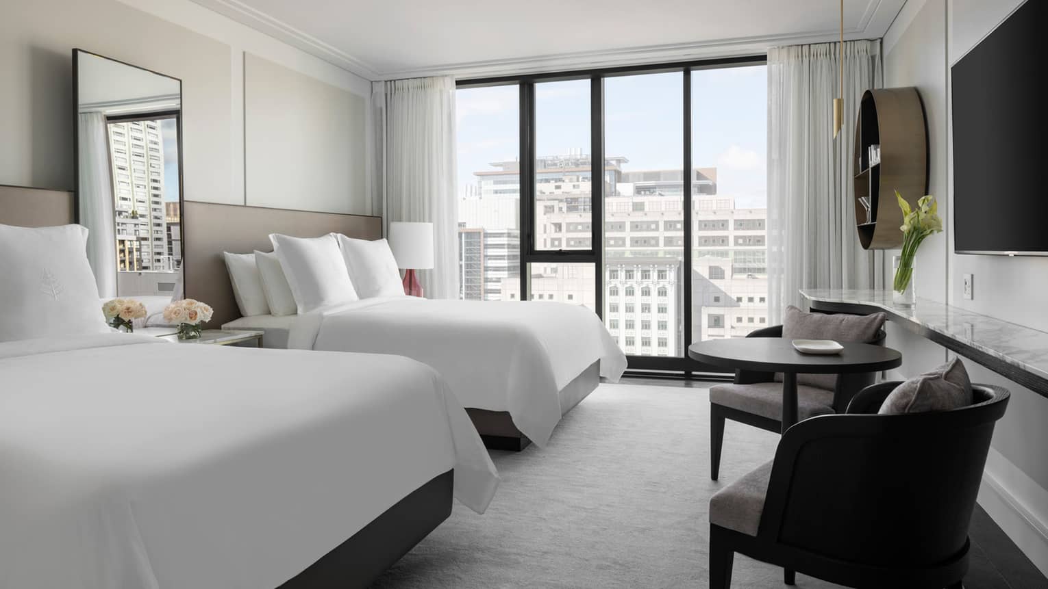 A bright hotel room with two double beds featuring white linens, large windows with city views, wall-mounted TV, and small seating area with table and chairs