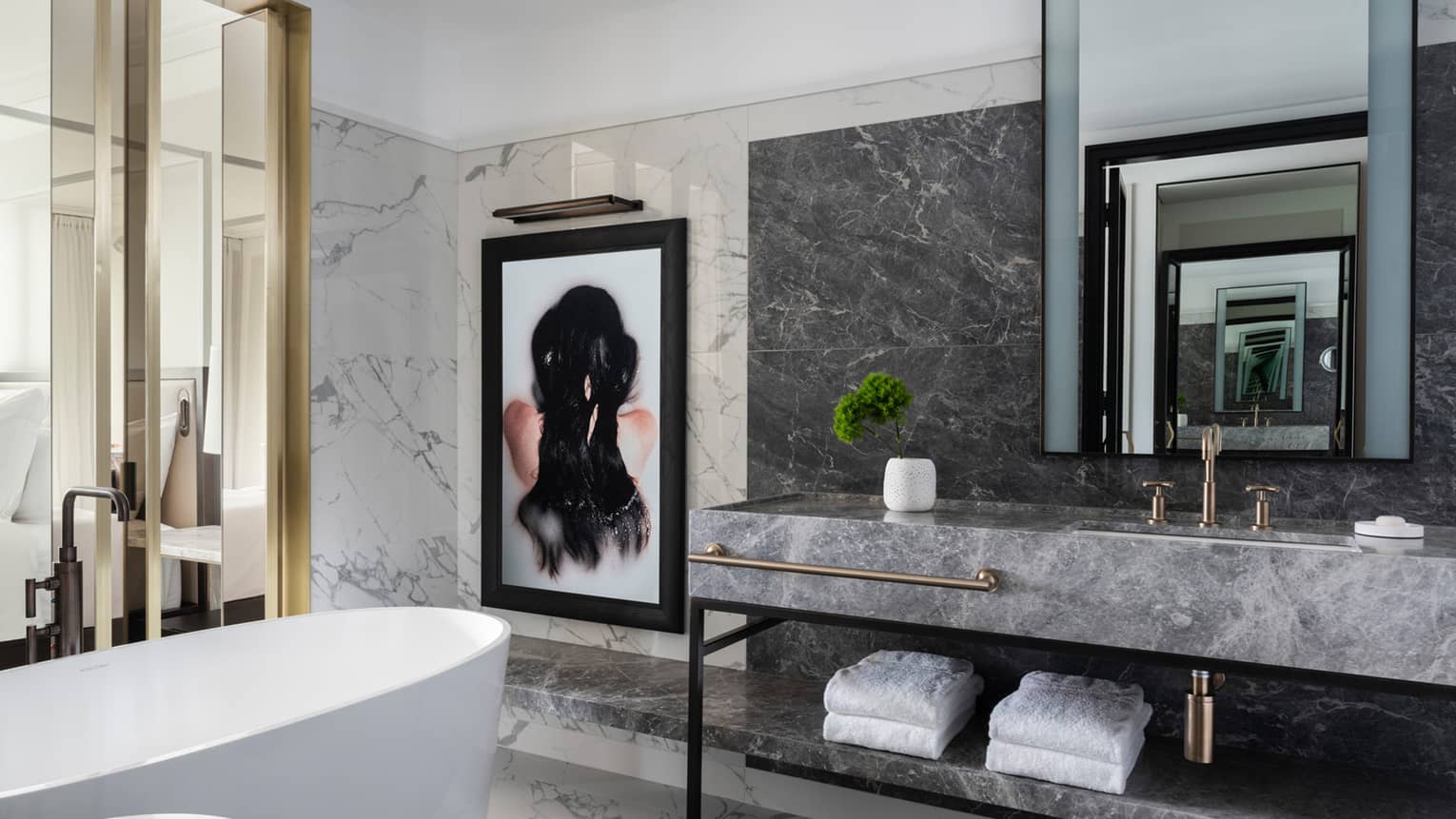 A freestanding tub, grey marble vanity and a framed work of art are set beside glass doors that open to a bedroom