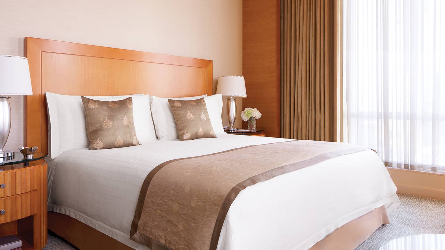 Embroidered accent pillows on hotel bed beside silver lamp, nightstand, flowers