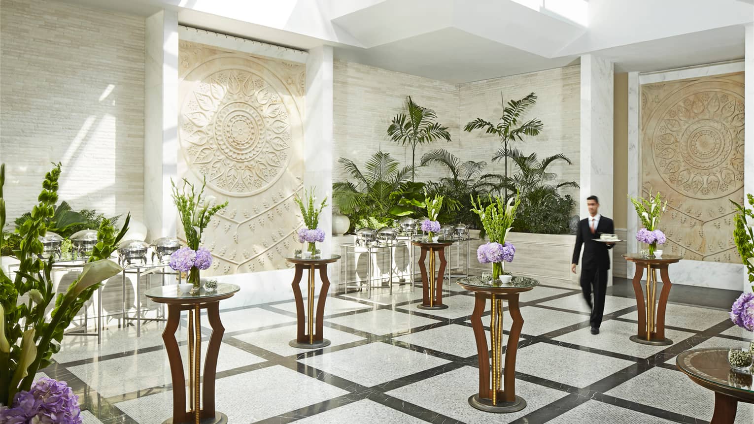 Server with tray walks through white marble room with accent tables, floral displays